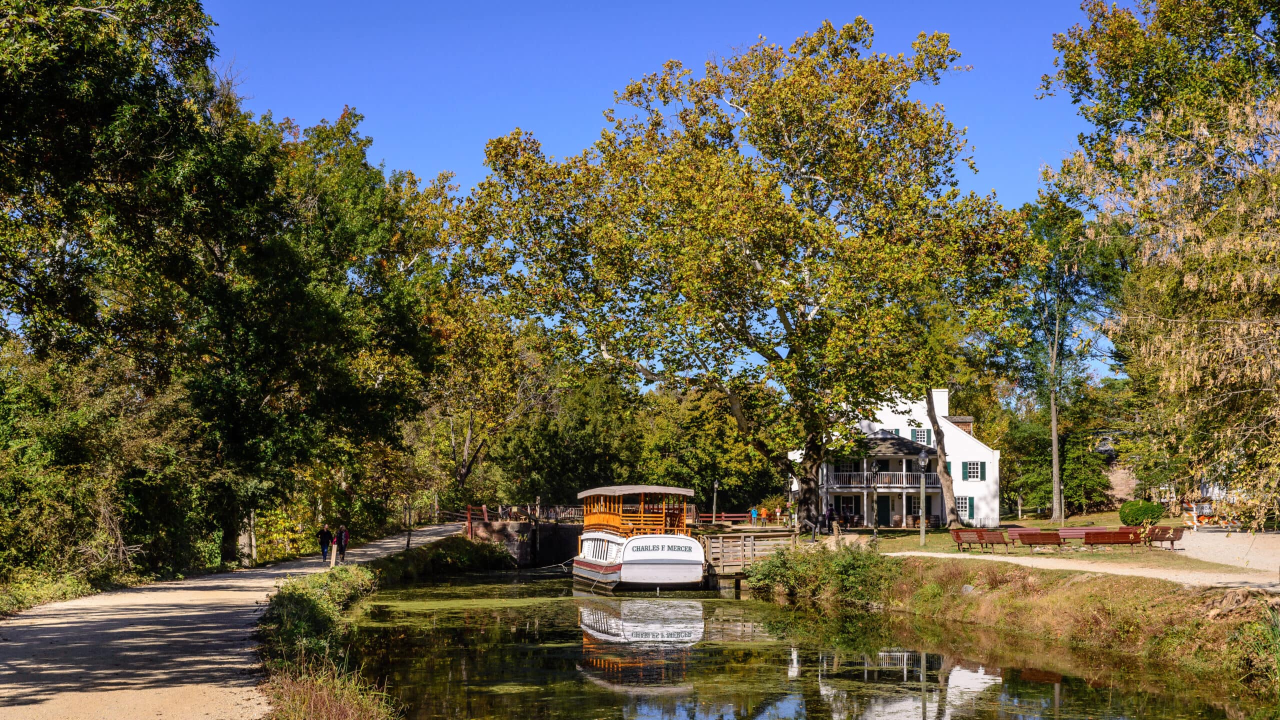 A landscape picture of the Great Falls Tavern Visitors Center and a boat in water. On the left side of the photo is a part of the towpath.