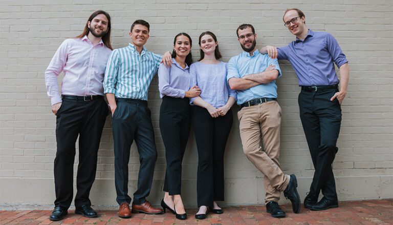 ars gratia populi group members in blue shirts and neutral pants against a white brick wall 768x439