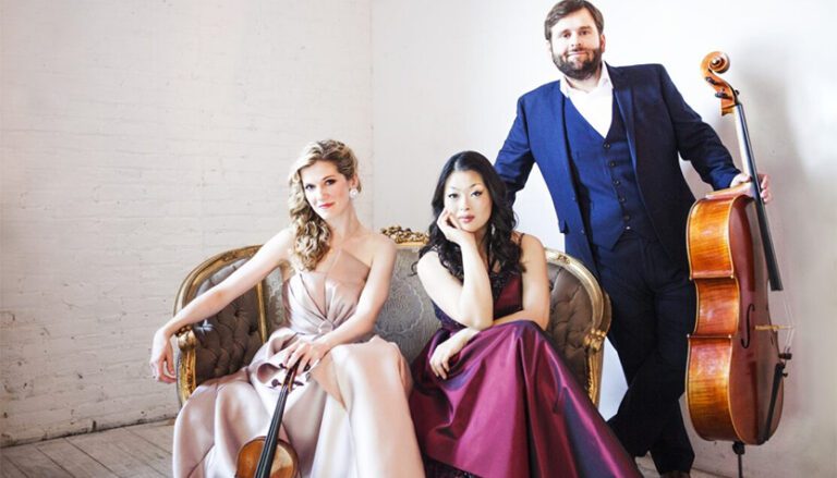 neave trio members posing on a fancy couch in front of a white brick wall 768x439